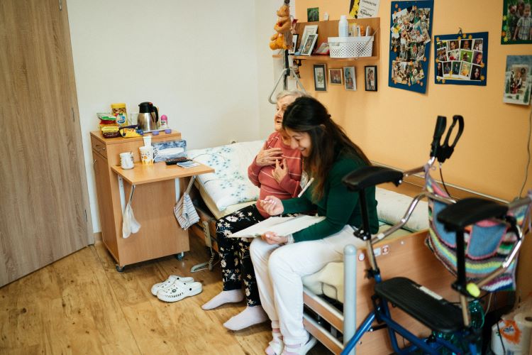 young woman sits with elderly woman on her bed in a room decorated with photos is a care facility