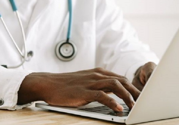 Physician of color's hands typing on laptop, wearing white lab coat and blue stethoscope