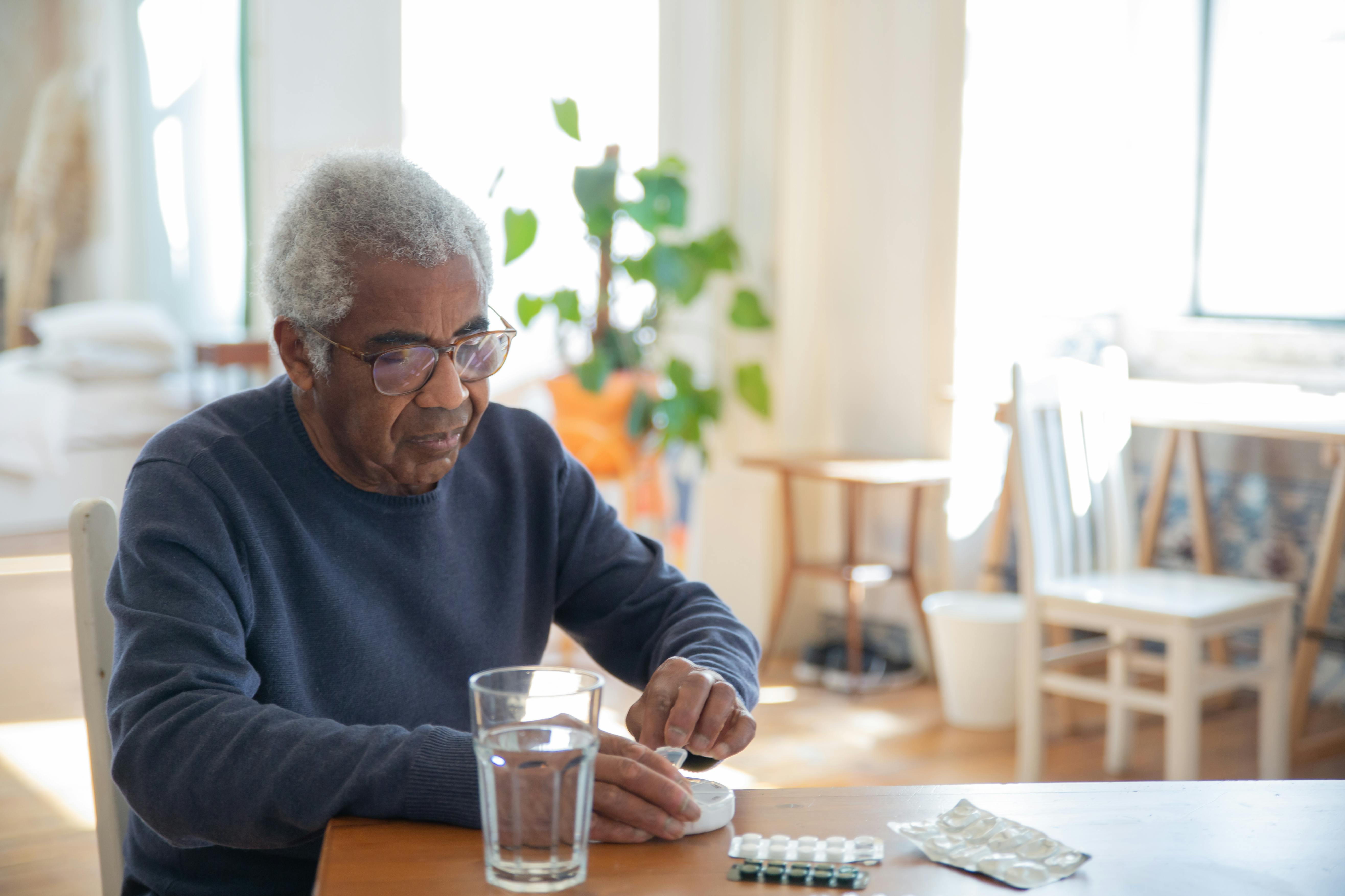 man sitting at table in a sunny room with glass of water and ready to take medication